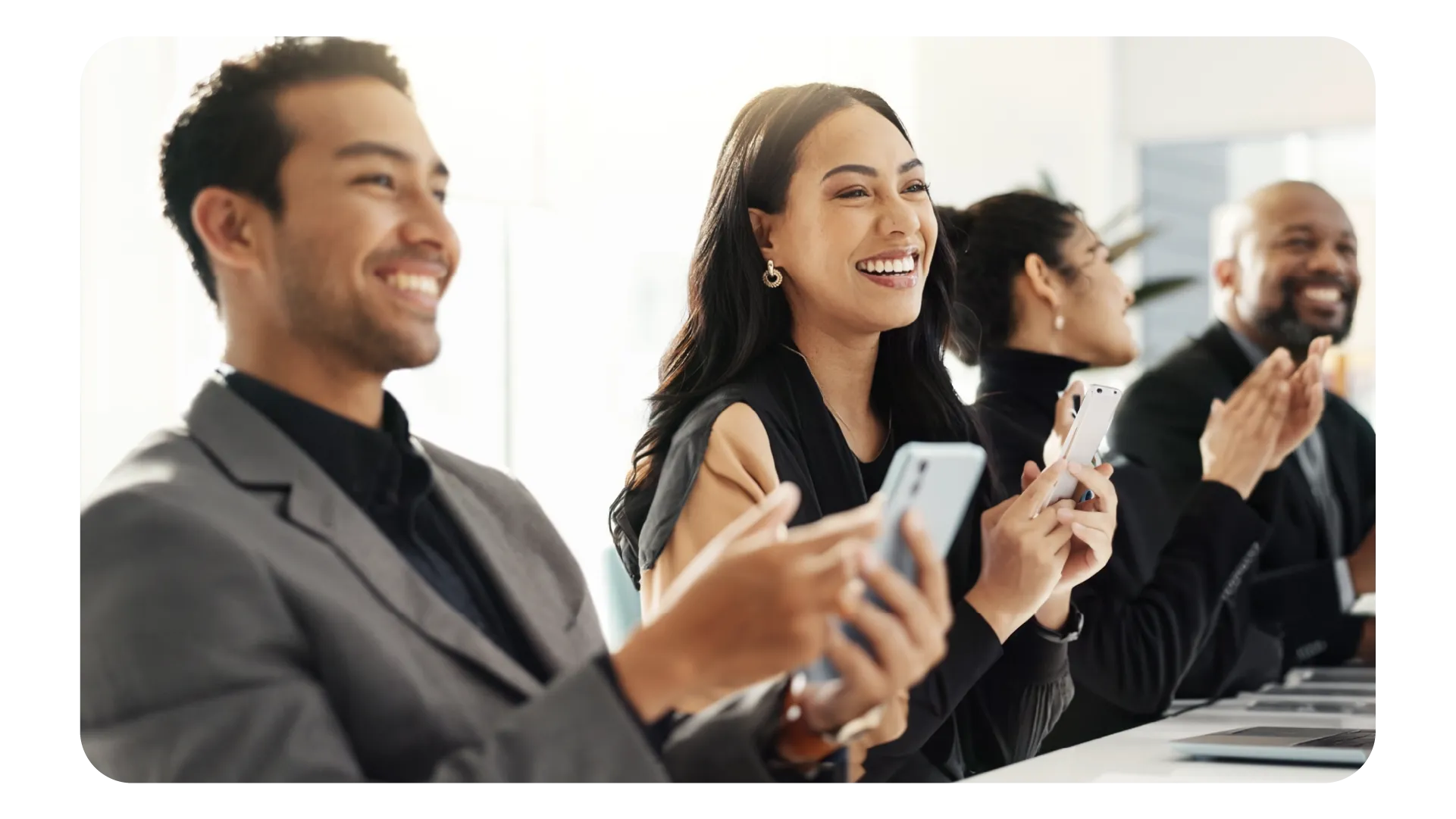 Four diverse professionals sitting in a row, smiling and clapping, holding smartphones in a bright office setting.