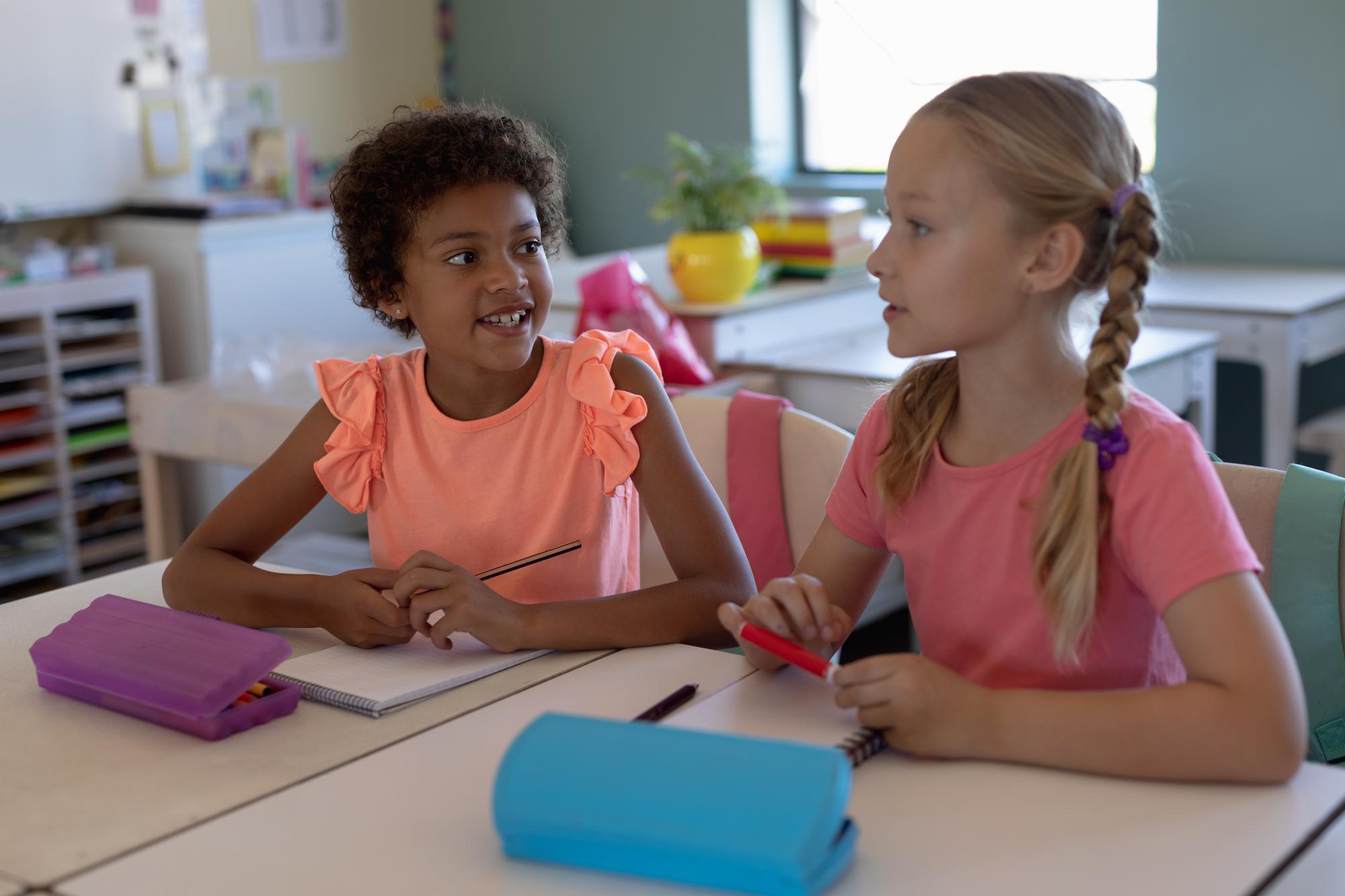 Two young schoolgirls sitting together at a classroom desk, talking and sharing ideas during a lesson