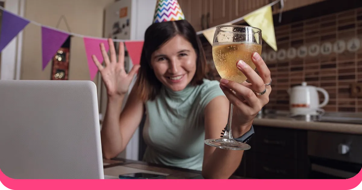 Woman in a party hat toasting friends over a video call from her kitchen