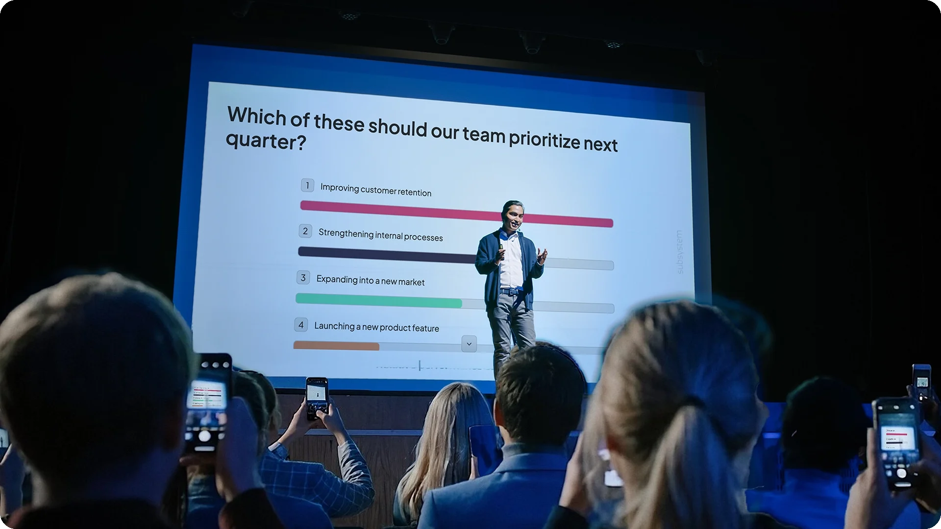 A speaker on stage in front of live ranking poll results, with the audience voting on their own phones